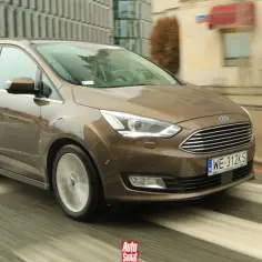 Brązowy Ford Grand C-Max w ruchu miejskim, z rozmytym tłem budynków i ulicy.