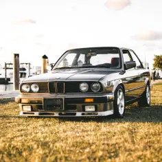 Klasyczne bmw e10 na trawie przy nabrzeżu, z łodziami w tle.
