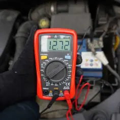 Ręka w czarnej rękawiczce trzyma miernik pokazujący 12.72V. W tle widać akumulator samochodowy, sugerując kontrolę napięcia.