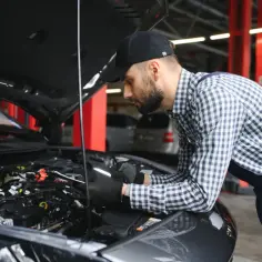 Mechanik w czapce i kratowanej koszuli pochyla się nad silnikiem samochodu, wykonując precyzyjny dolot do skomplikowanych przewodów.