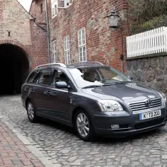 Szary Toyota Avensis T25 2.0 D4D na brukowanej uliczce, obok ceglanego budynku z łukiem.