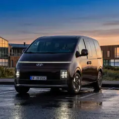 Ciemnobrązowy Hyundai Staria van zaparkowany na mokrym parkingu o zachodzie słońca.