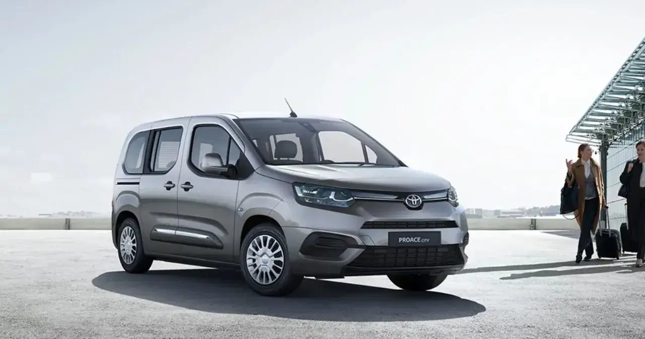 Srebrny Toyota Proace City Verso zaparkowany na zewnątrz, dwie kobiety z bagażami idą w jego kierunku.