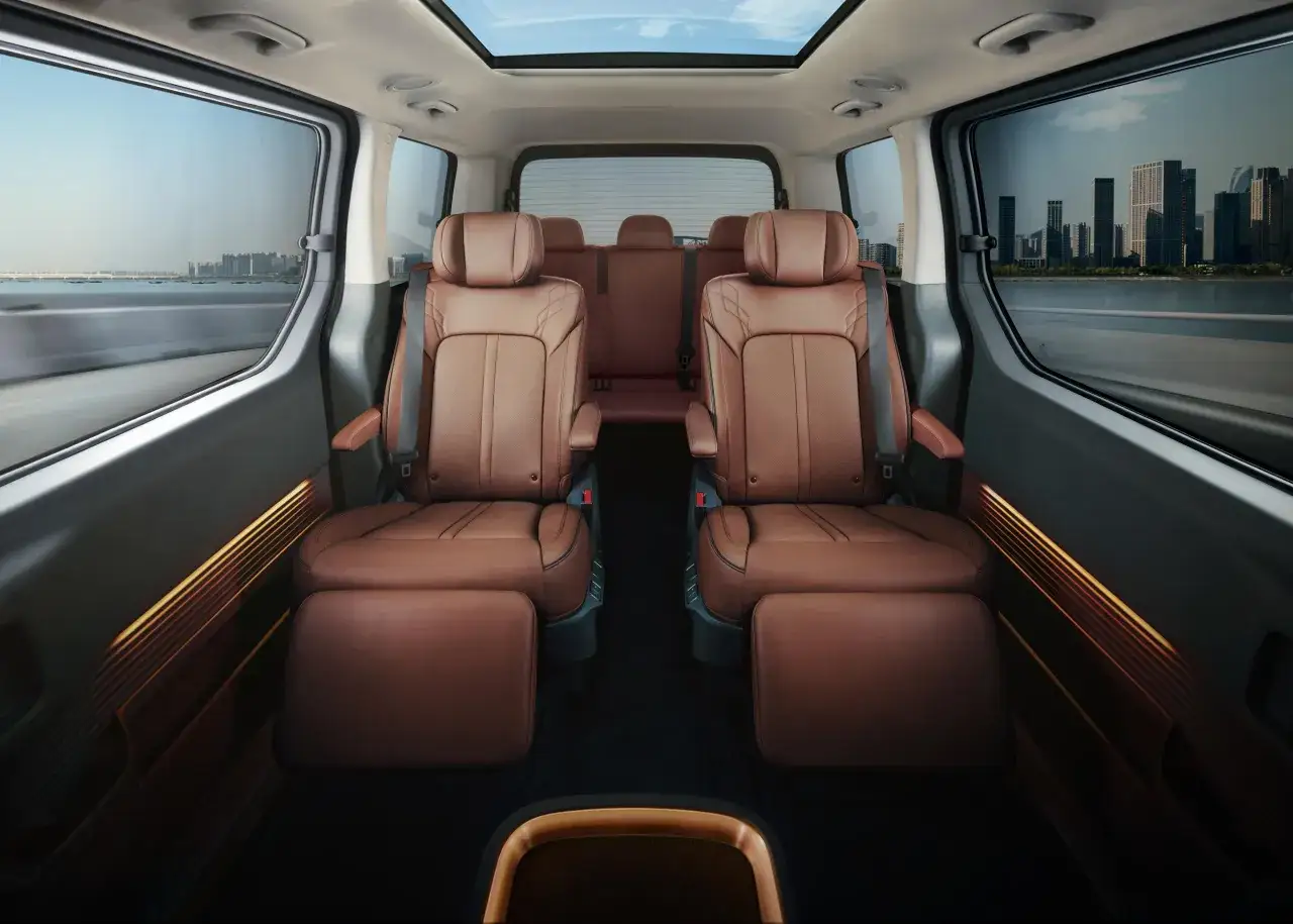 Wnętrze luksusowego hyundai van z brązowymi skórzanymi fotelami, podnóżkami i panoramicznym dachem. Widok na miasto przez okna.