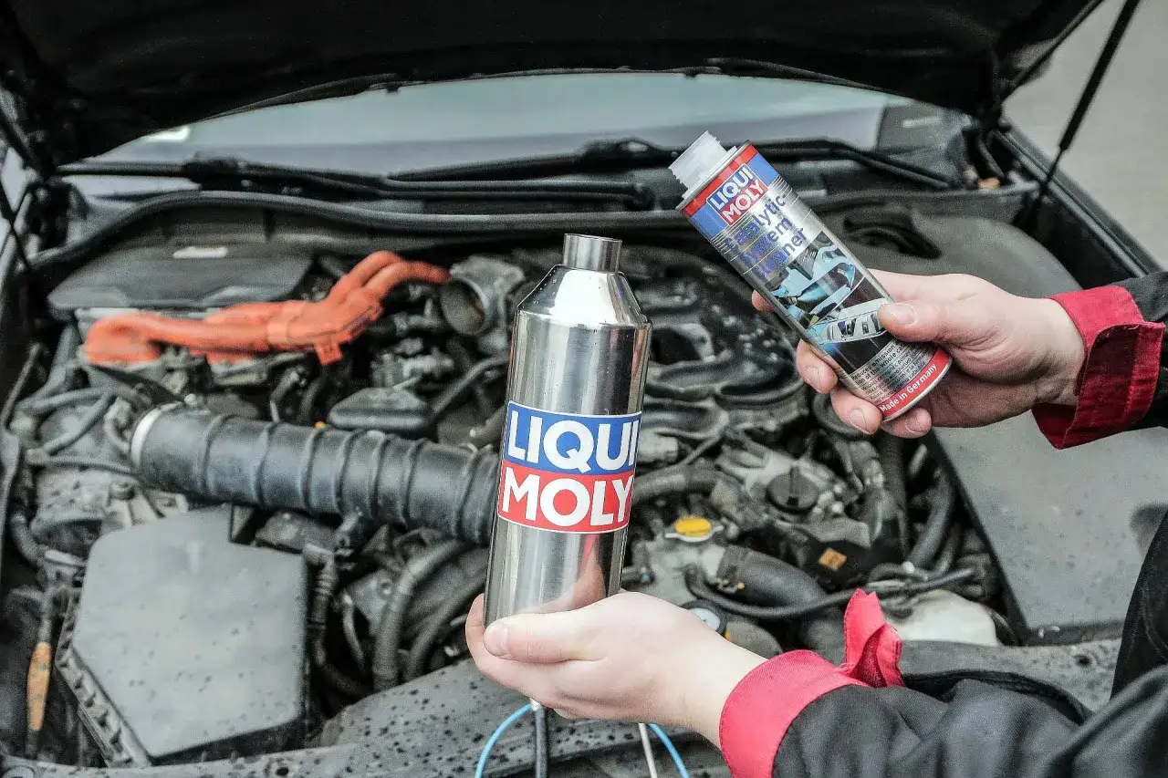 Ręce trzymające preparaty Liqui Moly do czyszczenia katalizatora na tle silnika samochodu.