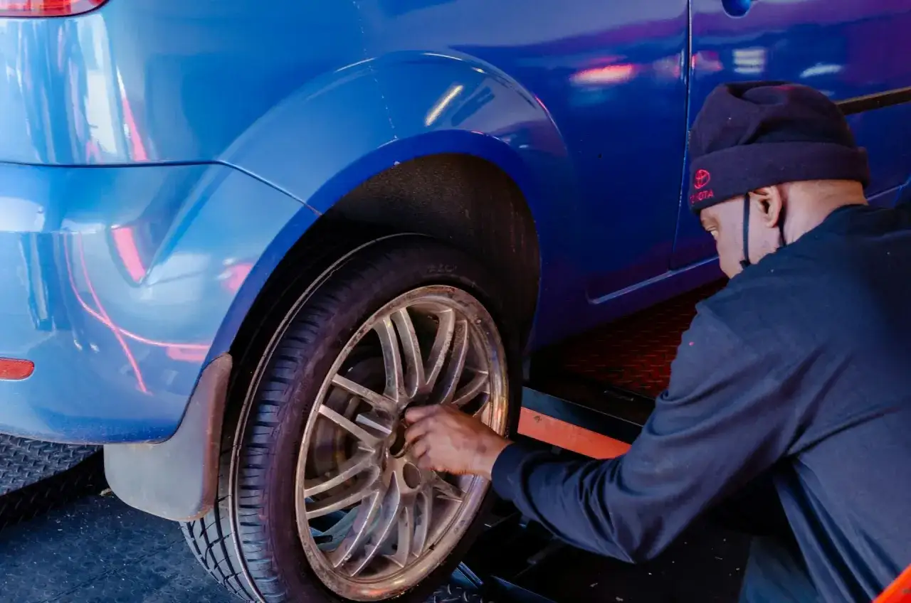Mężczyzna w czapce z logo Toyoty pracuje przy kole niebieskiego samochodu, pokazując, jak zmienić oponę.