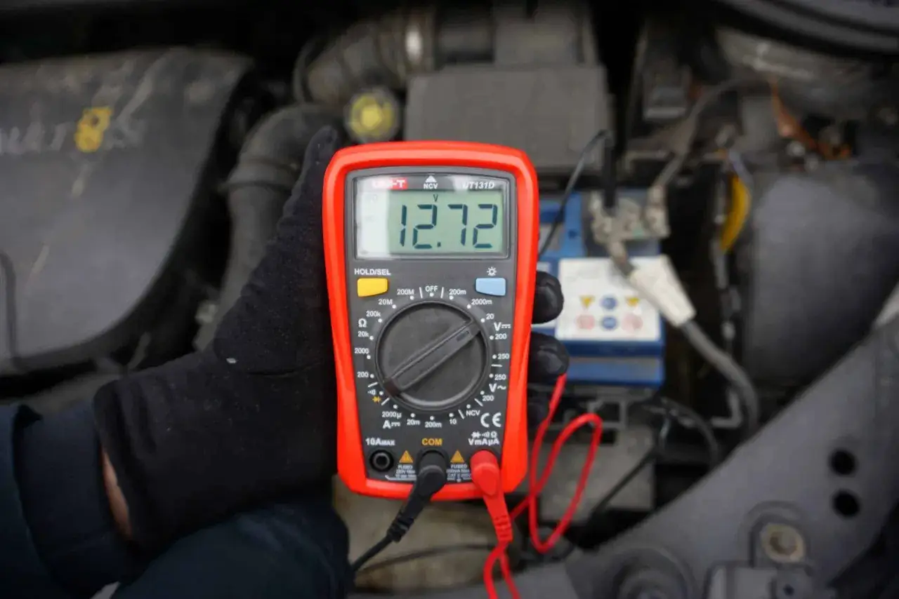 Ręka w czarnej rękawiczce trzyma miernik pokazujący 12.72V. W tle widać akumulator samochodowy, co sugeruje sprawdzanie napięcia.