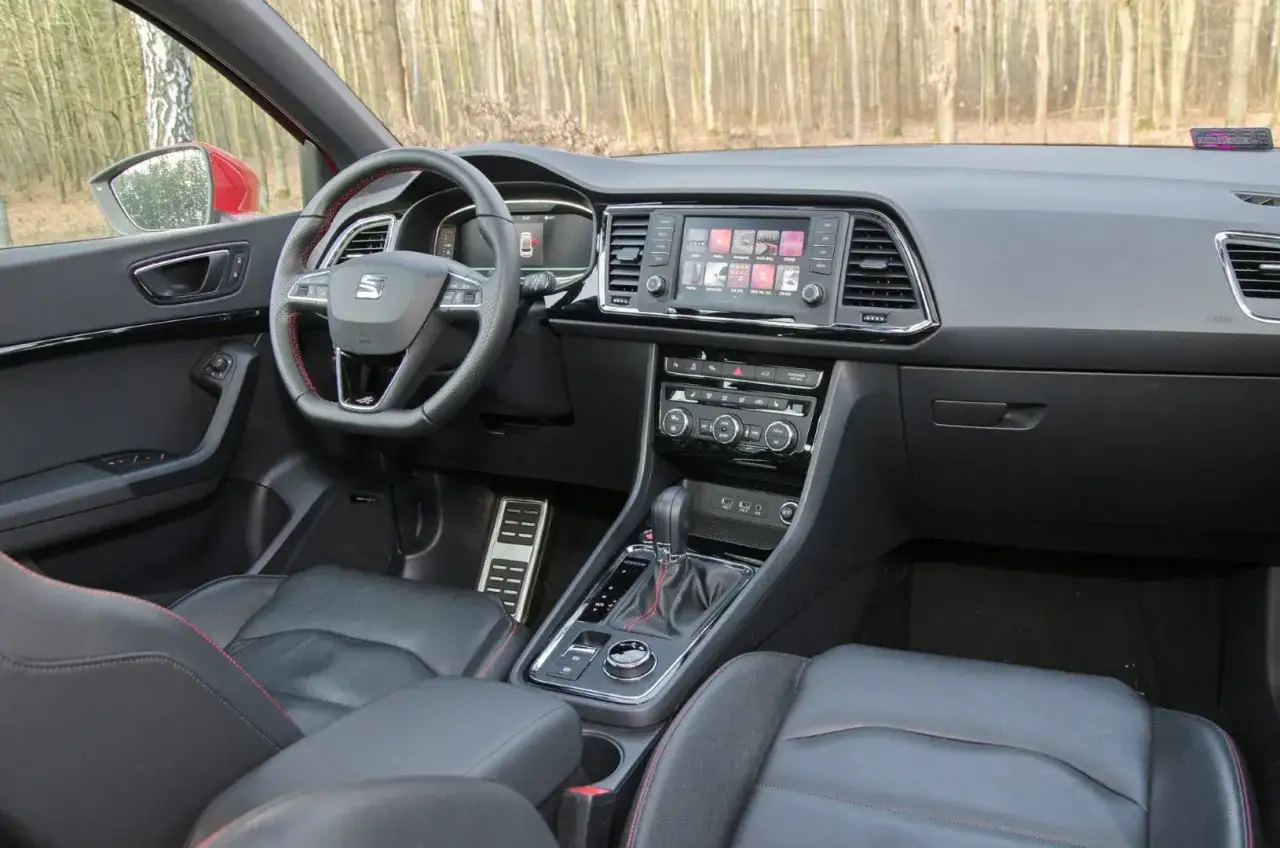 Wnętrze Seat Ateca FR z czarną tapicerką i czerwonymi przeszyciami. Widoczny ekran multimedialny, kierownica i dźwignia zmiany biegów.